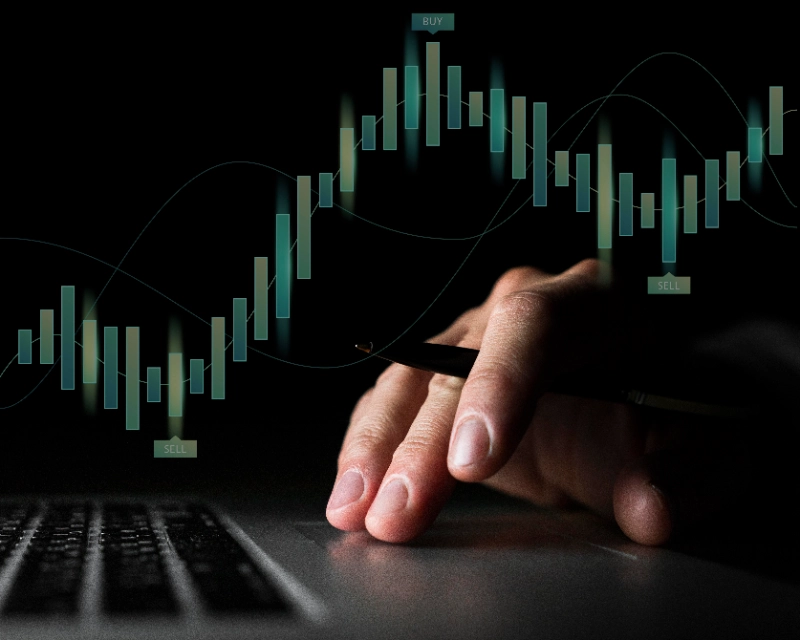 Mano usando una laptop en la oscuridad frente a un gráfico financiero digital interactivo con barras de velas y señales de compra y venta.