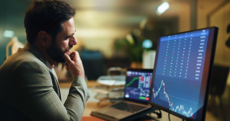 Un hombre de negocios reflexivo analiza gráficos e información bursátil en el monitor de su computadora durante la noche.