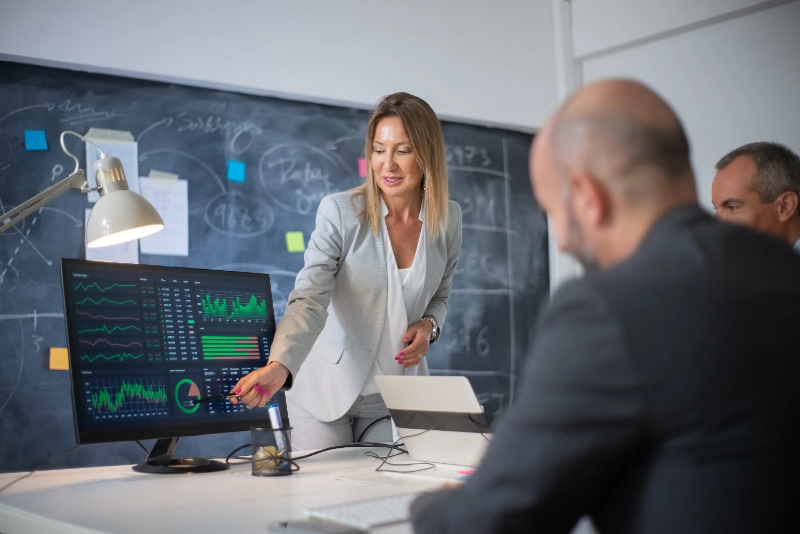 Mujer de negocios señalando gráficos de rendimiento en un monitor durante una reunión de trabajo con sus colegas.