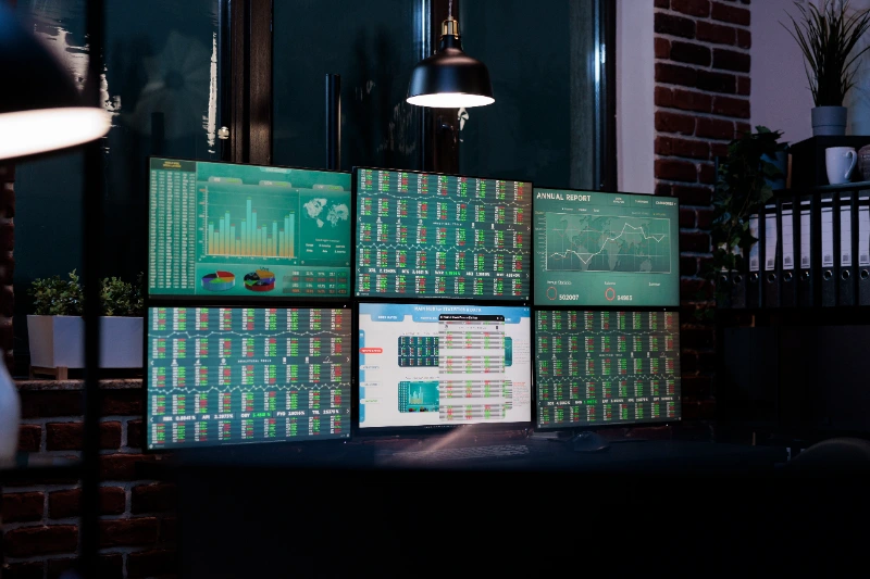 Estación de trabajo vacía en una oficina moderna iluminada con seis monitores que muestran datos bursátiles y gráficos financieros en tiempo real.