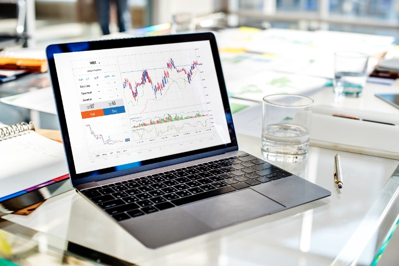 Laptop sobre una mesa de cristal mostrando gráficos financieros y de operaciones Forex.