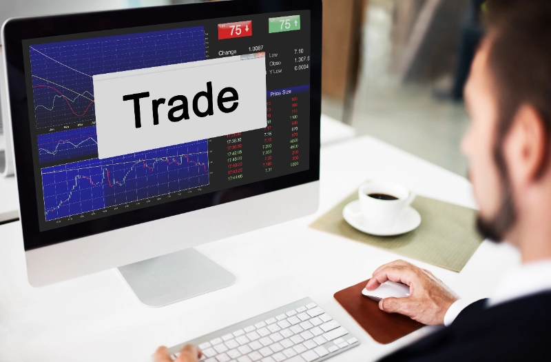 Hombre en traje frente a una computadora analizando gráficos bursátiles con la palabra Trade destacada en la pantalla.
