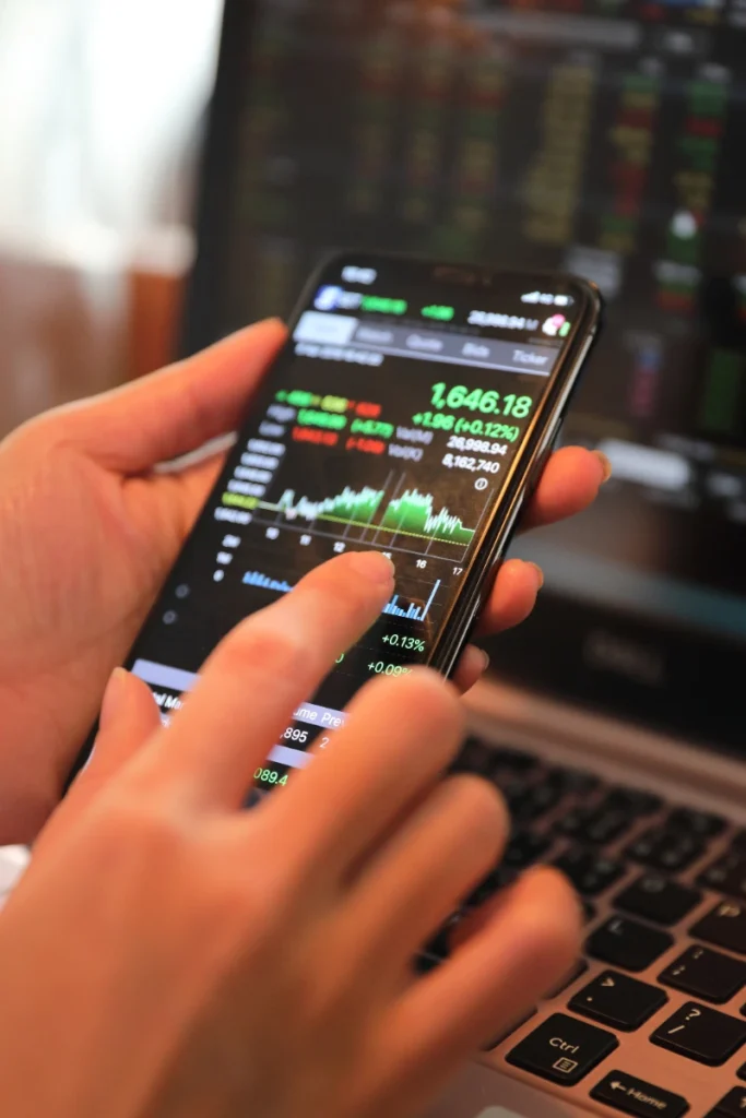 Persona revisando el gráfico bursátil y los datos financieros de un valor en la pantalla de su teléfono móvil frente a una computadora portátil.