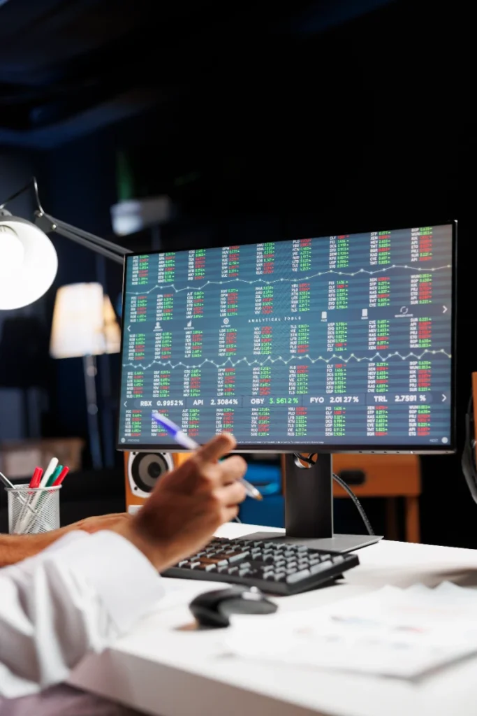 Persona sosteniendo un bolígrafo frente a un monitor que muestra tablas de datos bursátiles y paneles financieros en un entorno de oficina.