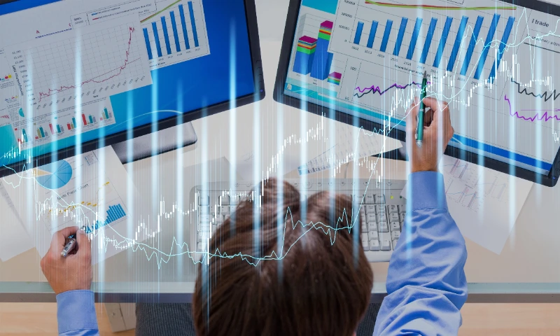 Vista superior de un analista financiero trabajando con dos monitores y documentos impresos, con superposición de datos bursátiles holográficos.