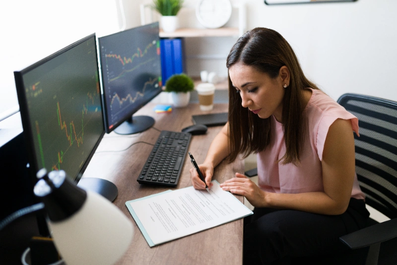 Mujer profesional firmando un contrato financiero en su escritorio junto a dos monitores que muestran gráficos bursátiles.