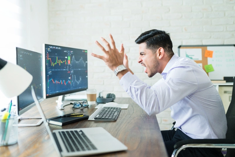 Un operador financiero frustrado grita frente a múltiples monitores que muestran gráficos bursátiles en su espacio de trabajo.