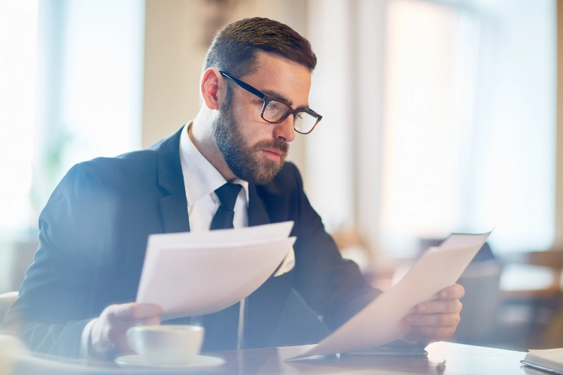 Un ejecutivo con gafas revisa atentamente varios documentos de papel en una oficina o cafetería bien iluminada.