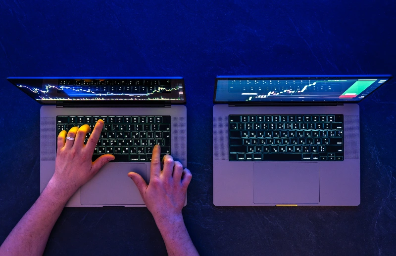 Vista superior de las manos de un operador tecleando en uno de dos portátiles abiertos con gráficos bursátiles en directo sobre un fondo oscuro e iluminación azul.