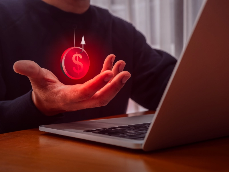 Una persona frente a un ordenador portátil sostiene una moneda roja brillante enganchada en un anzuelo virtual, simbolizando un ciberataque de phishing.