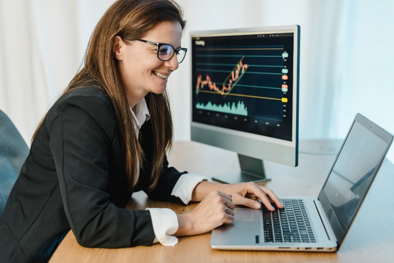 Una broker sonriente en traje formal opera en línea con un portátil y un monitor que muestra gráficos financieros.