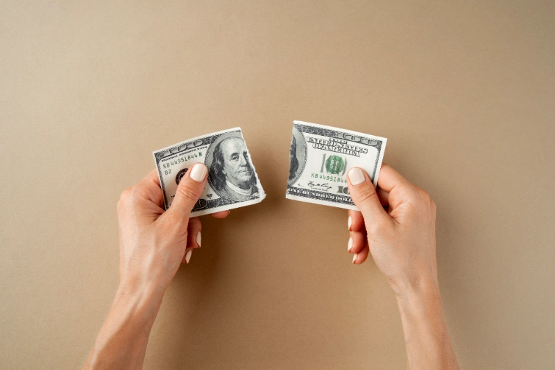Vista superior de dos manos sosteniendo las mitades separadas de un billete de 100 dólares rasgado sobre un fondo beige, simbolizando pérdida financiera o división económica.