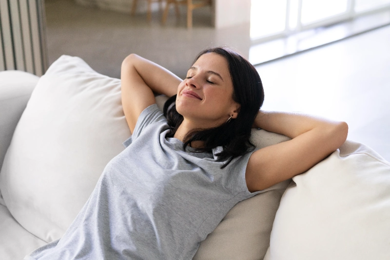 Una mujer descansa relajada en un sofá blanco con las manos detrás de la cabeza y los ojos cerrados.